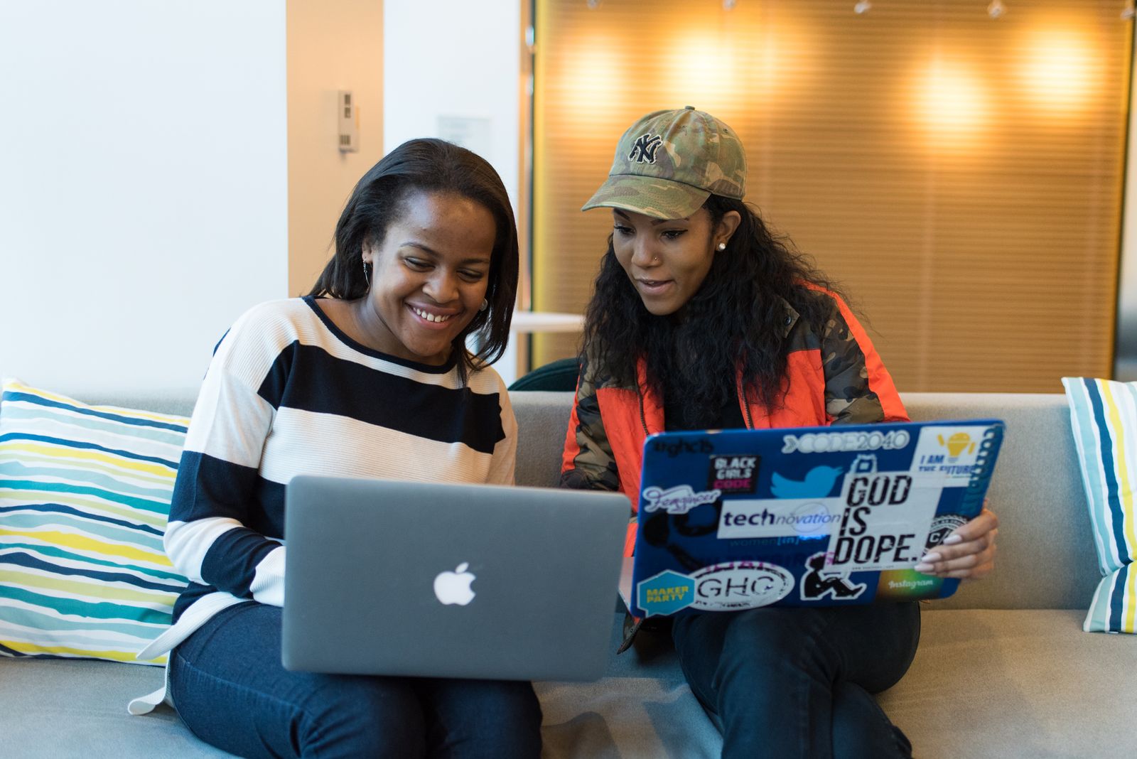 Two colleagues on a sofa, smiling as they look over work on their laptops together.